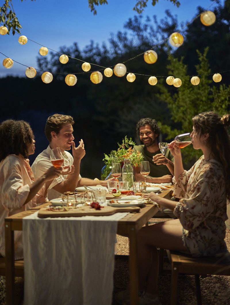 Cuatro amigos disfrutan de una cena al aire libre bajo luces cálidas. El ambiente es jovial, con vino y comida en la mesa de madera.