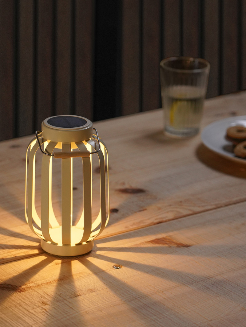 Una linterna solar blanca ilumina una mesa de madera. Un vaso de agua y galletas crean un ambiente tranquilo y acogedor al anochecer.