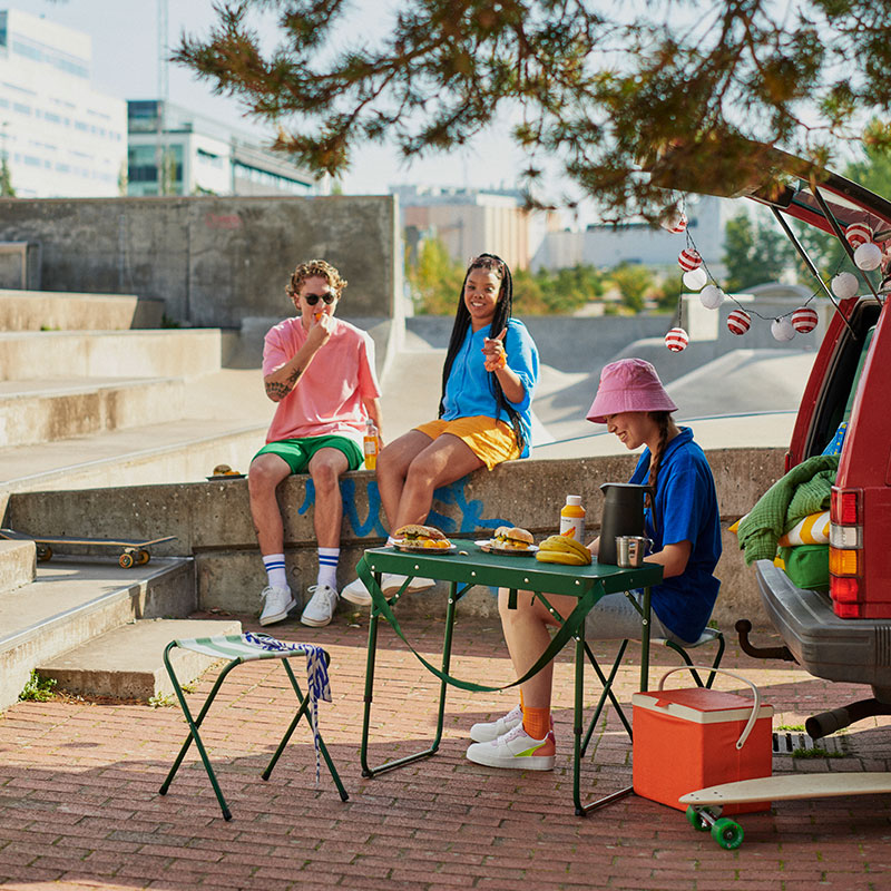 Young people sharing food at a skate park. Green table, orange cooler, relaxed and sunny atmosphere.