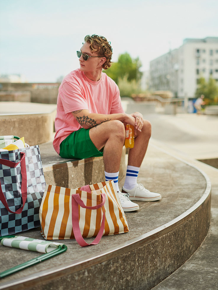  Young man in pink T-shirt and green shorts, sitting outdoors with patterned bags. Urban and casual vibe