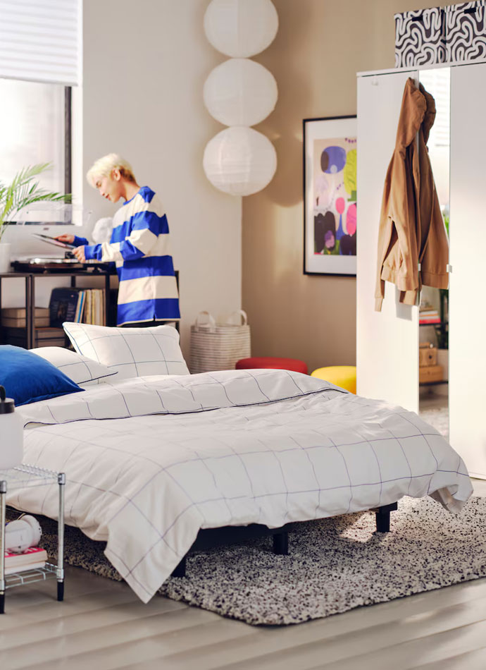 A large bed in a student bedroom, there is a mirrored dresser with a light brown jacket hanging in the background and a young man playing music on a record player.