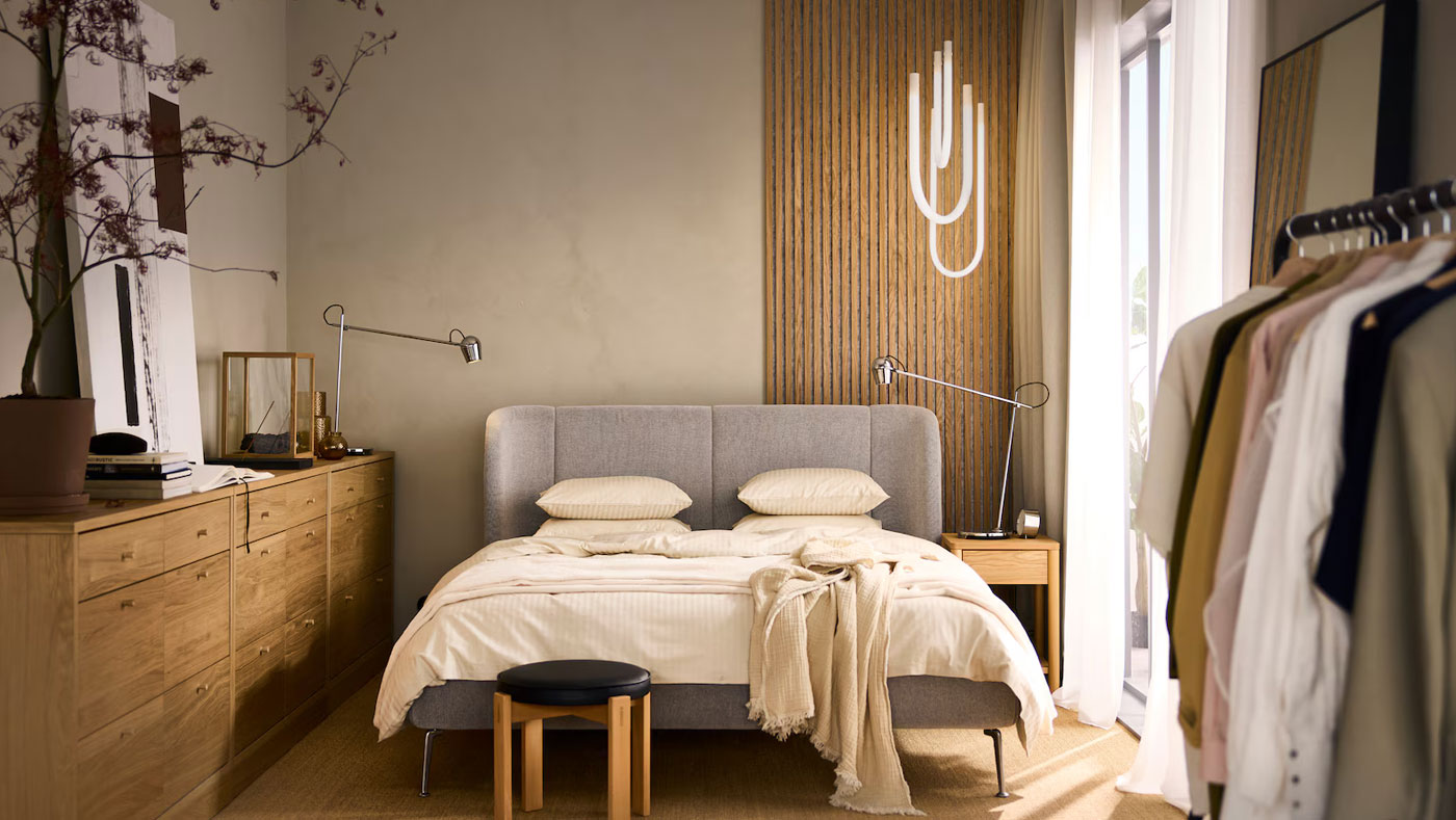 Modern bedroom with gray bed, wooden dresser, clothes rack, and warm accents. Cozy atmosphere.