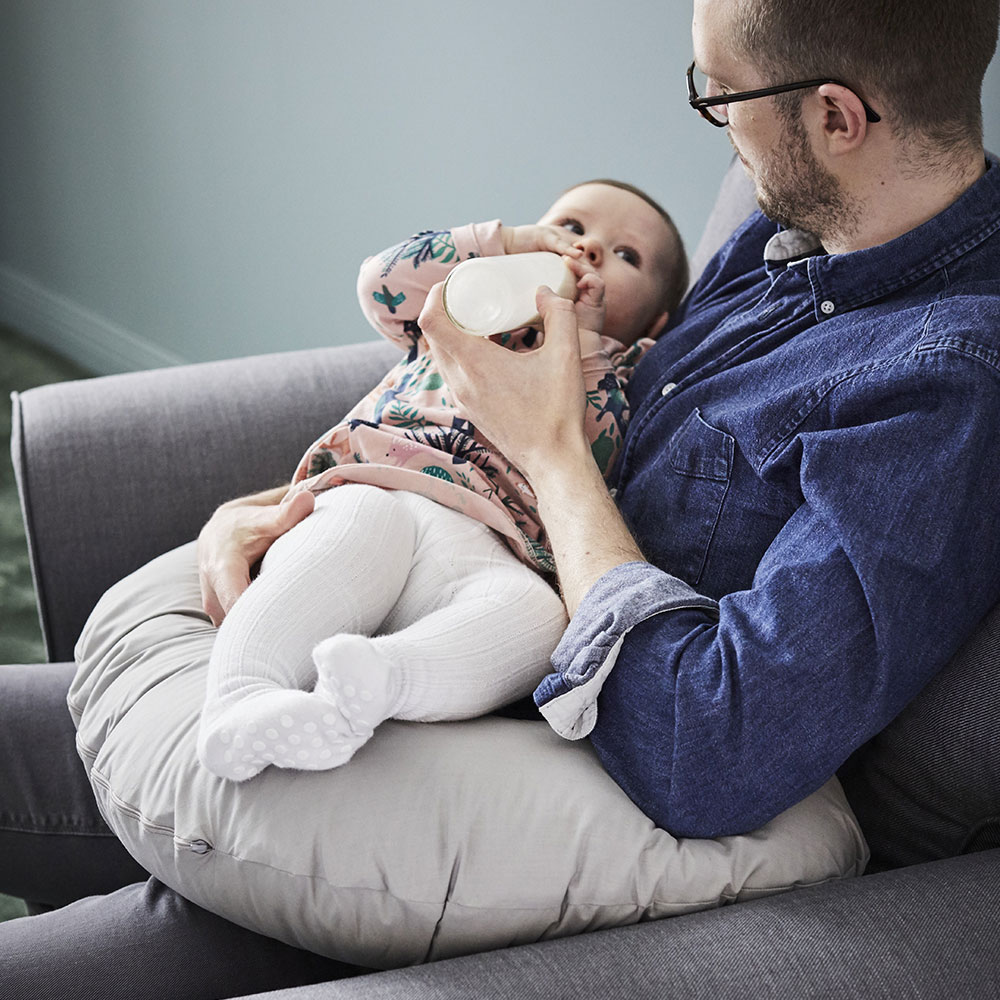 A man feeding milk to a baby while sitting in a STRANDMON armchair with a LEN nursing pillow.