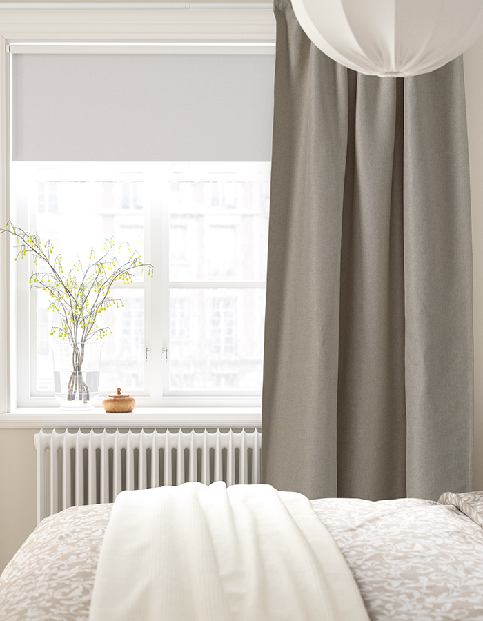 Bright bedroom with bed dressed in light tones, thick grey curtain, white roller blind, and a vase with decorative branches on the windowsill.