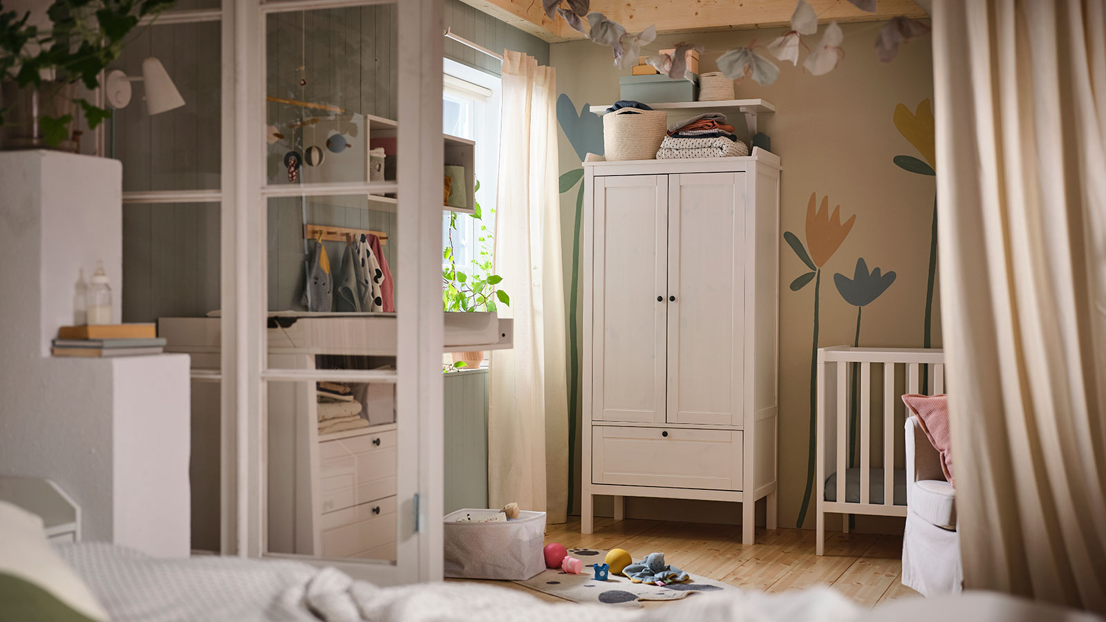A baby's room, a changing table in front of a window, a modular shelf, a closet next to a crib and toys on the floor next to a curtain.