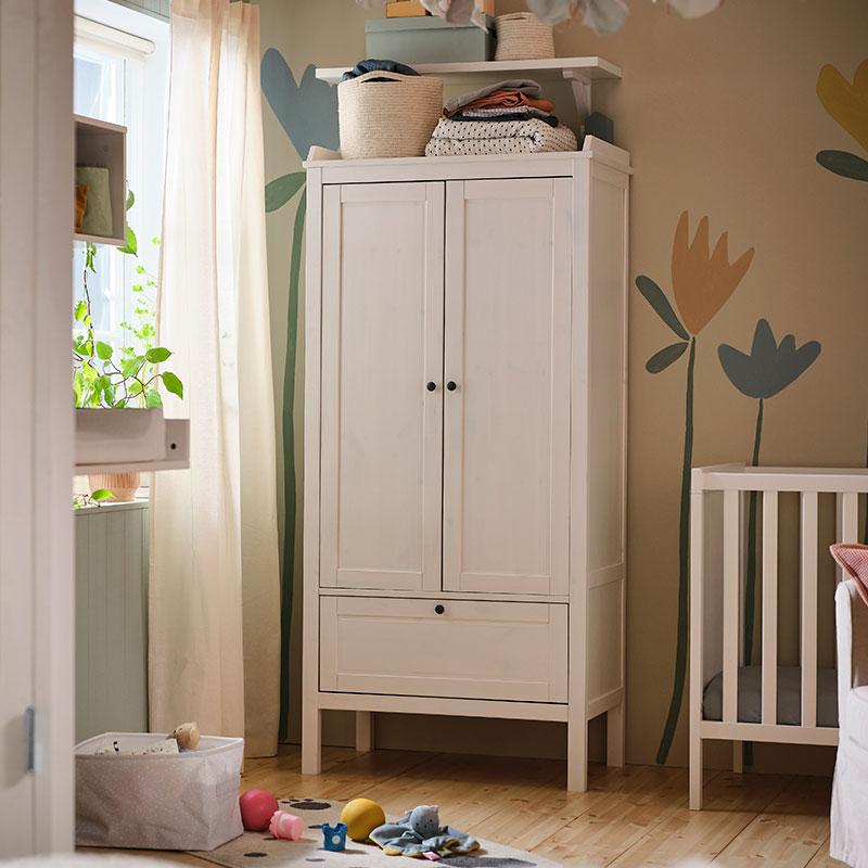 A baby's room, a changing table in front of a window, a modular shelf, a closet next to a crib and toys on the floor next to a curtain.