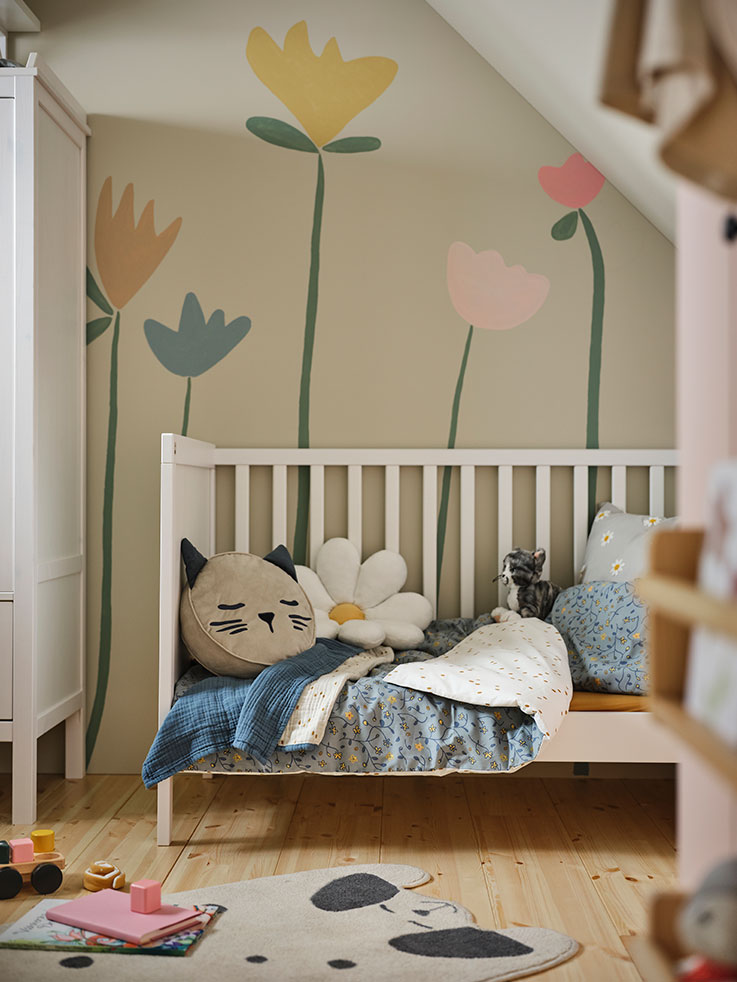 A crib filled with cushions in the shape of flowers and kittens and quilts, toys on the floor.
