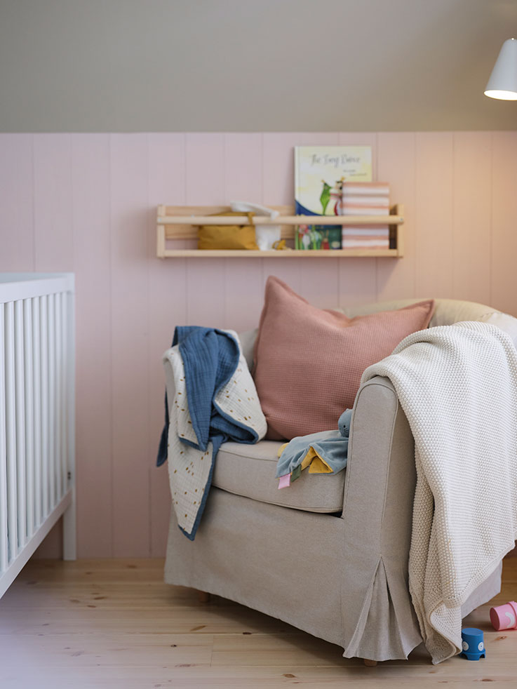 Armchair with textile cover with a pink cushion and a quilt and a baby blanket, there is a hanging pine shelf on the wall, and part of a cradle can be seen.