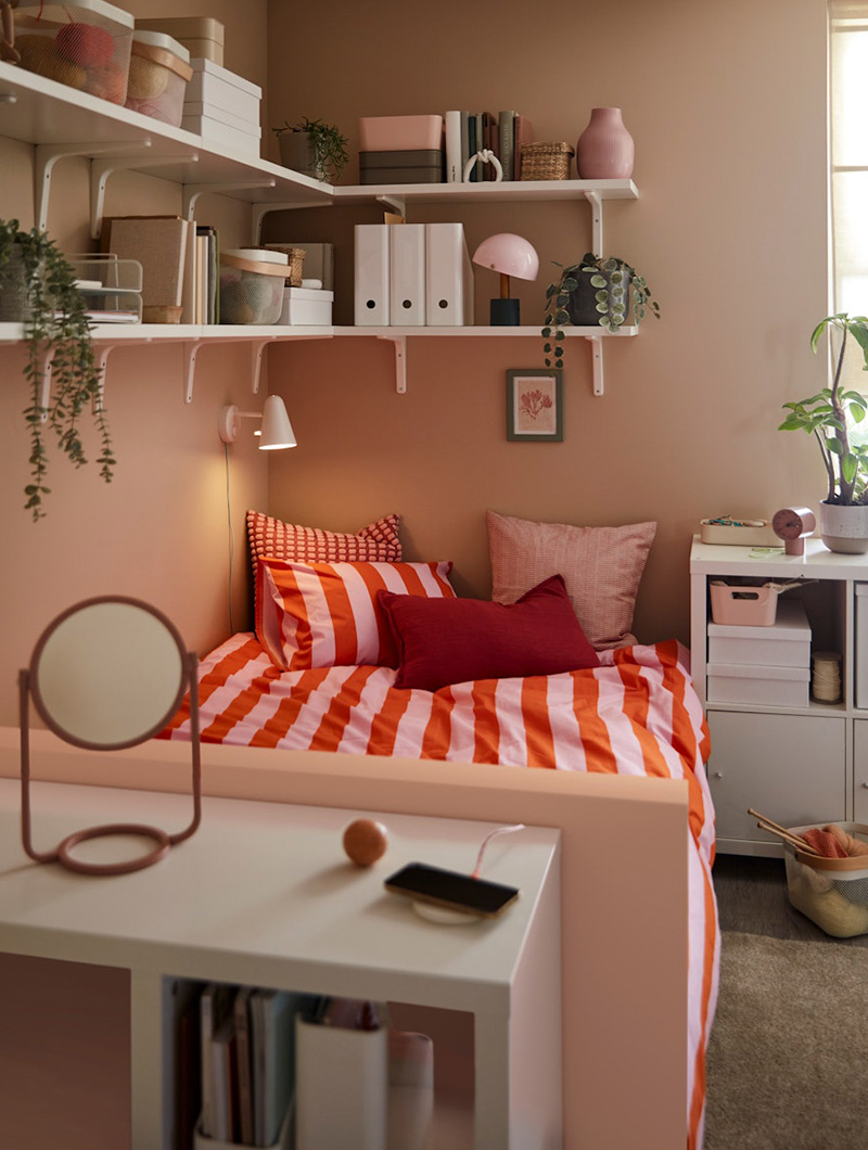  Bedroom in pink and red tones, striped bed, white shelves, and mirror. Youthful ambiance.