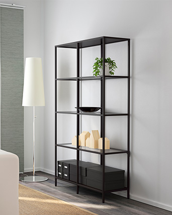 A tall, black metal shelving unit holds a green plant, a black bowl, wooden house models, and black storage boxes. A white floor lamp stands left.