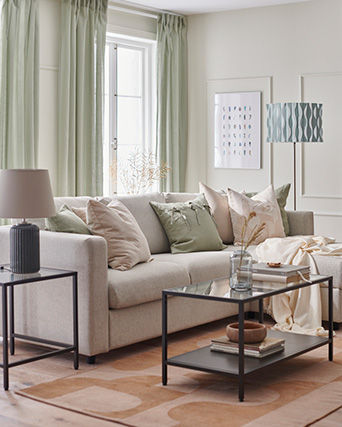Beige sofa, varied cushions, dark tables, black lamp, blue patterned floor lamp. Light green curtains and decor fill the room.