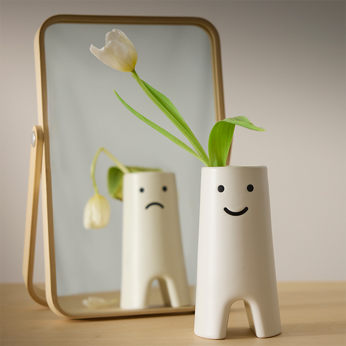 Florero blanco con rostro sonriente reflejado en un espejo pequeño