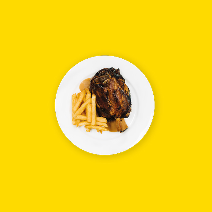 Plato blanco con codillo de cerdo asado y patatas fritas sobre un fondo amarillo.