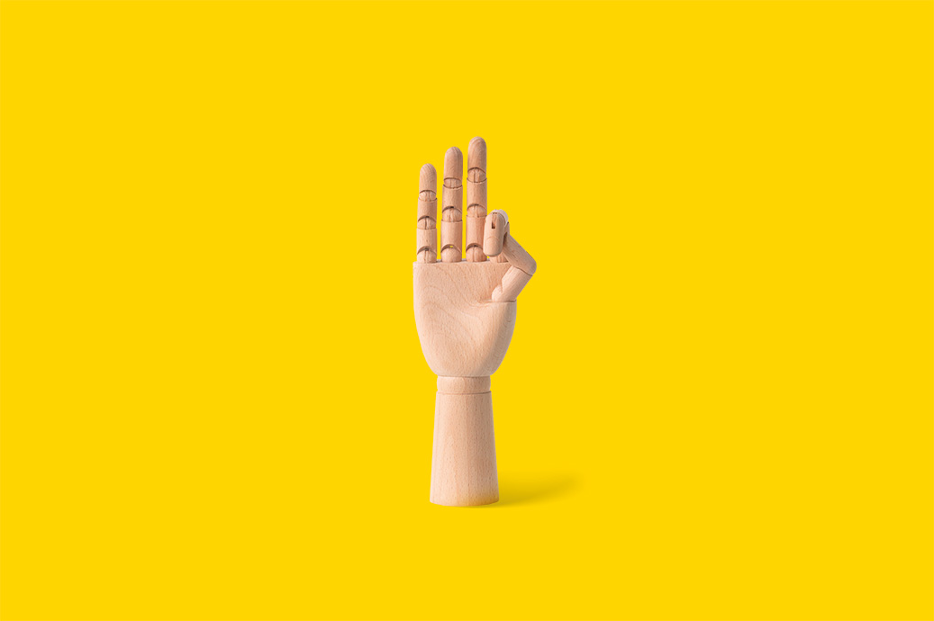 Mano de madera articulada mostrando tres dedos levantados sobre fondo amarillo.