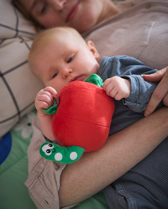 Bebé sujetando un peluche con forma de manzana roja de la que asoma un gusanito verde.