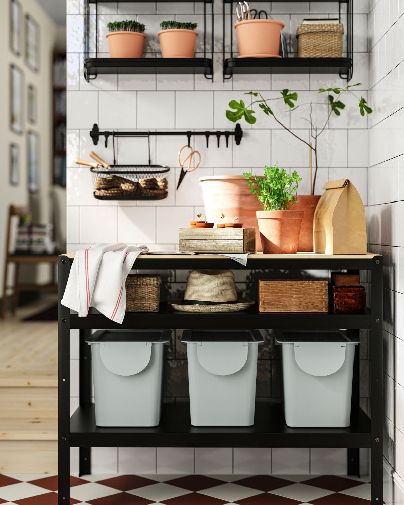 Functional utility area with black shelving, potted plants, and gray recycling bins on a tiled wall.