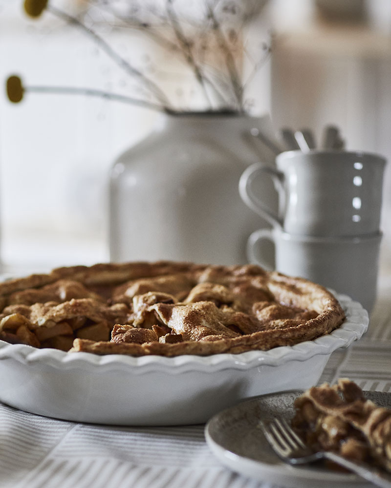 An off -white HÖSTAGILLE pie dish filled with apple pie sits on a stripey tablecloth, surrounded by cutlery and vases.