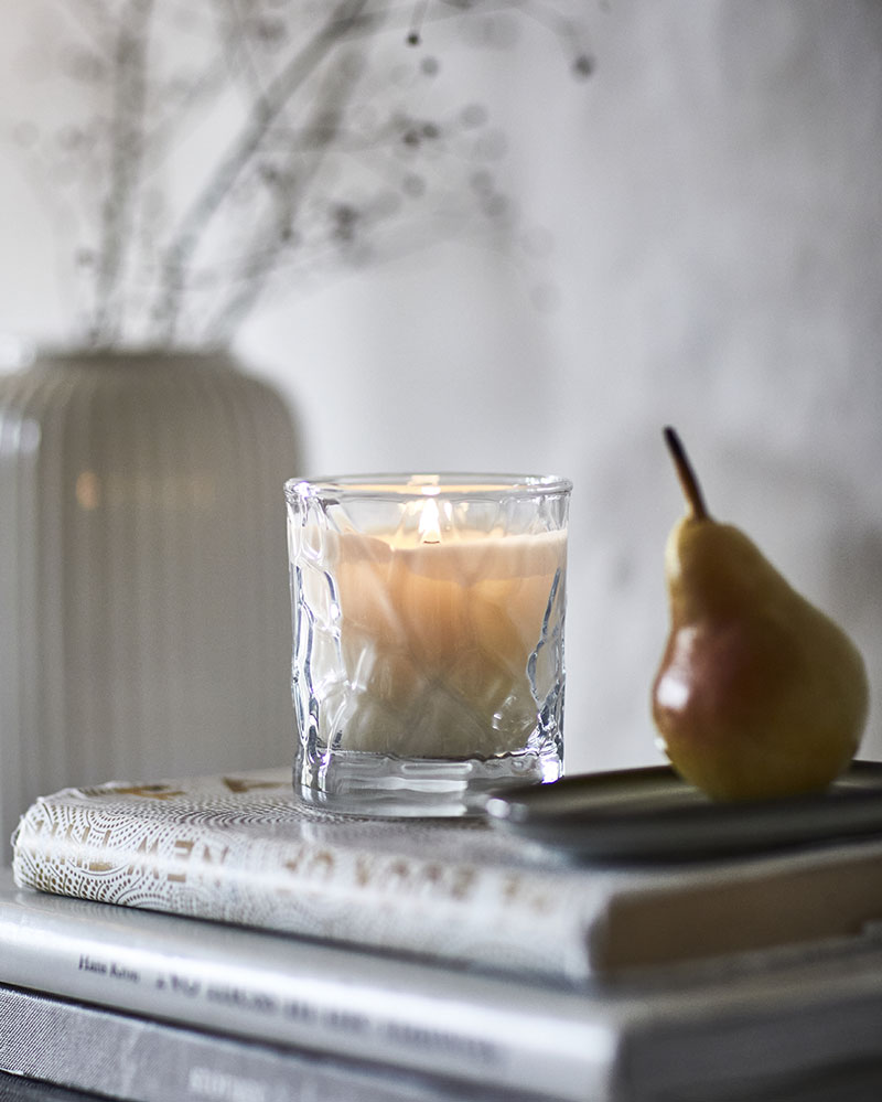 A scented HÖSTAGILLE candle in a glass jar is placed on stacked books beside a decorative vase and a pear