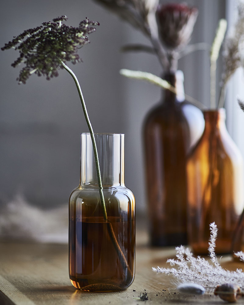 A brown HÖSTAGILLE vase holding a dried fl ower, elegantly placed on a wooden table with soft ambient light