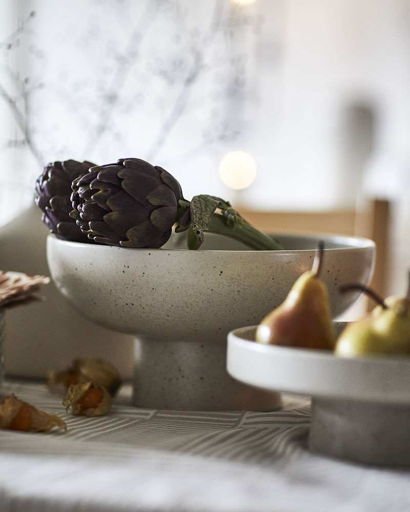 A light beige HÖSTAGILLE serving bowl holds artichokes on a table, next to a HÖSTAGILLE serving stand with pears in it.