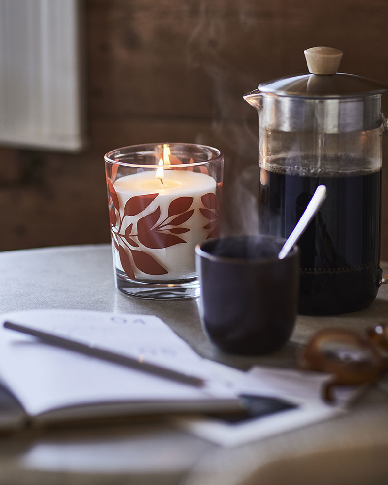 A HÖSTAGILLE scented candle in a glass sits on a table beside a French press and a steaming mug.