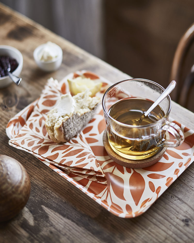 A HÖSTAGILLE tray with an orange pattern displays a slice of buttered bread, a glass of tea, and a HÖSTAGILLE paper napkin