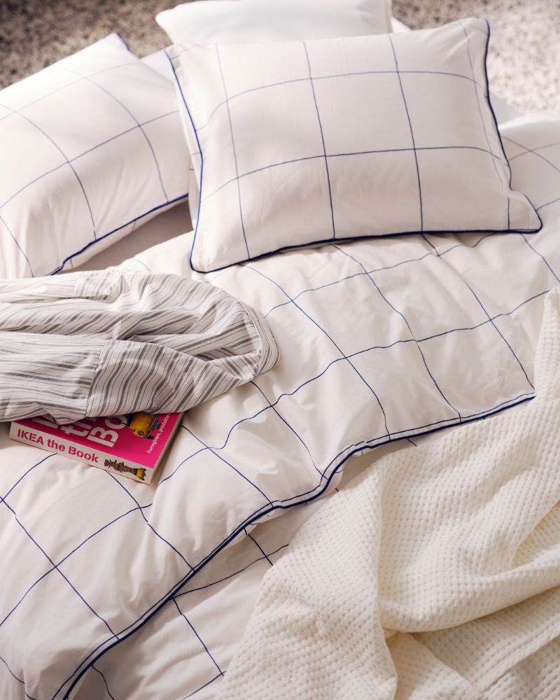 White-and-blue-checked BREDVECKLARE bed linen, a white waffle blanket and pink-andwhite book on a bed.