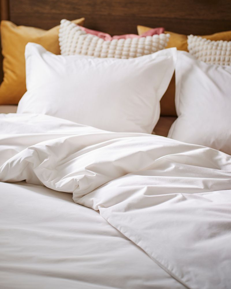 A bed made up with HAMPFLOCKEL bed linen with the duvet folded back and bright colour cushions behind the pillows.