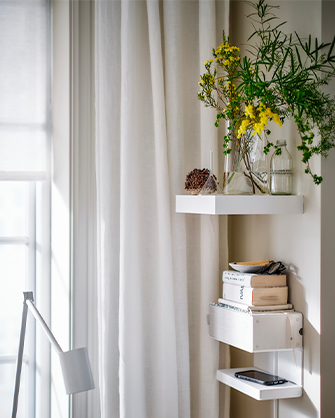 Rincón de una habitación con estanterías flotantes blancas y cortinas claras. Las estanterías sostienen jarrones con plantas, libros y un teléfono, organizando el espacio vertical.