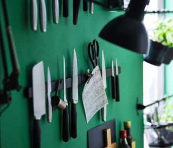 A green wall with diff erent kinds of knives, a pair of scissors, and more attached to a black HULTARP magnet knife rack. 