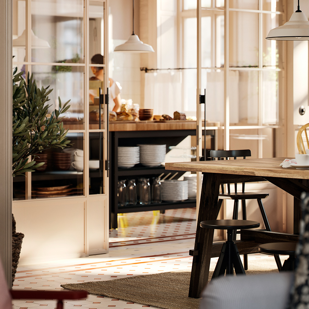 Part of an acacia SKOGSTA dining table with black legs is visible by a breakfast buffet area that is behind a glass wall.