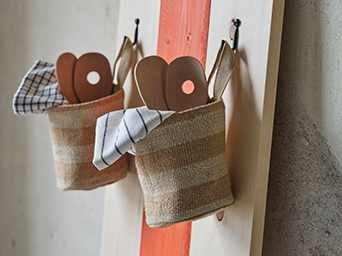 Dos cestas de almacenamiento MÄVINN de yute, llenas de utensilios de madera y paños de cocina, cuelgan por sus asas de unos ganchos en la pared.
