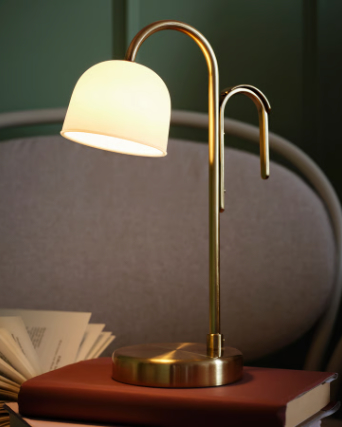 Una lámpara portátil LED TVÅMASTAD de color blanco ópalo descansa con elegancia sobre una pila de libros en un cómodo sillón.