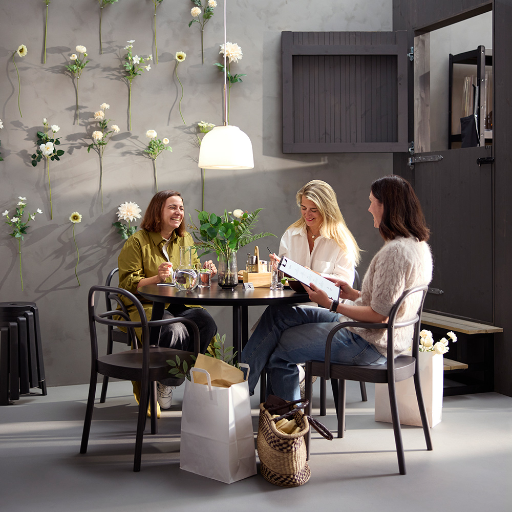 Café scene with three people at a black LISABO table, dark brown STOCKHOLM 2025 chairs, and a flower wall in the background.