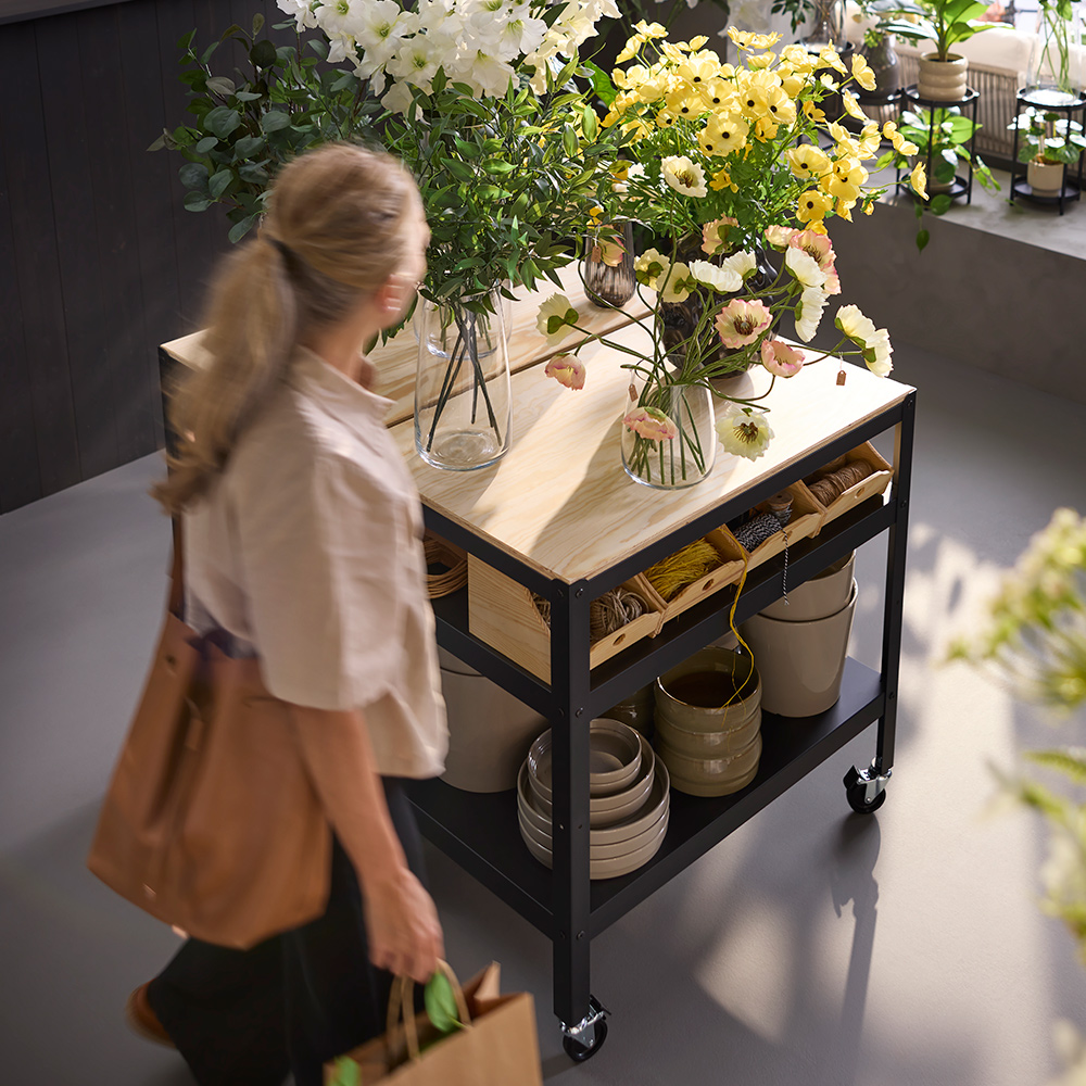 A person in a flower shop scene walking by a black BROR trolley made from pine plywood, holding plants and flowers.