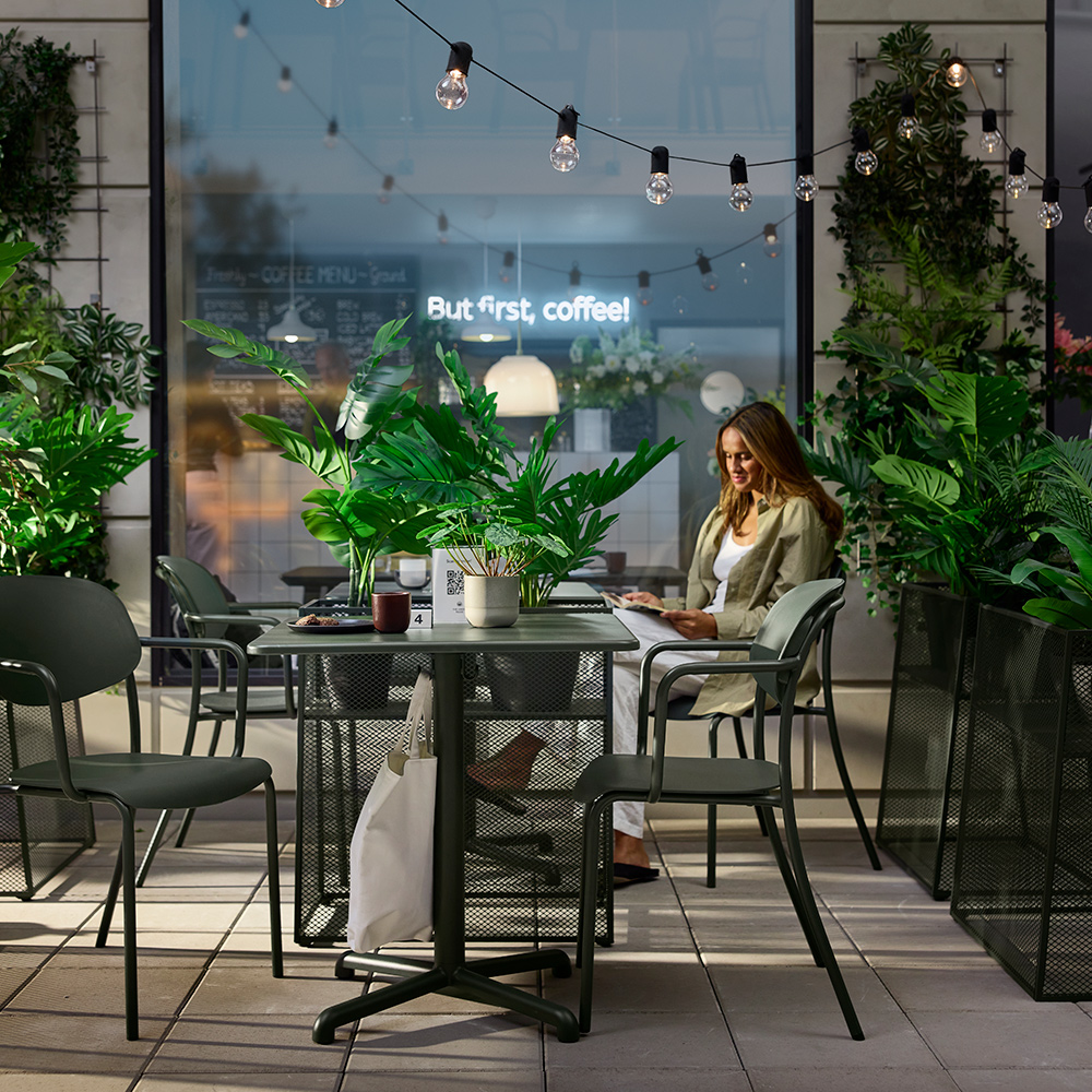 Café serving area outdoors featuring deep green VISINGSÖ tables, metal chairs and a person reading.
