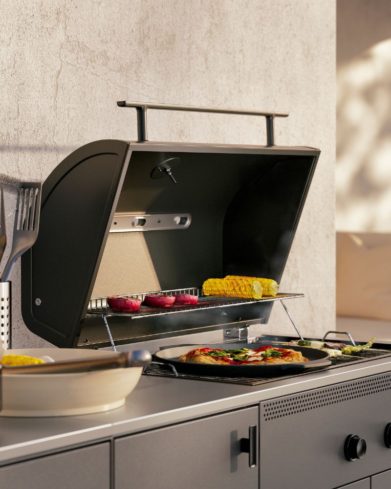 A close-up of the barbecue part of a dark grey BÅTSKÄR outdoor kitchen. The lid is open, showing a pizza and vegetables.
