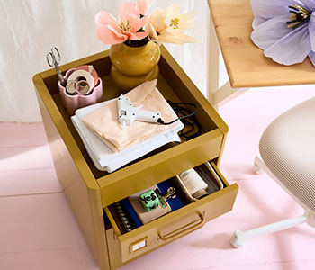 Mueble cajonero IDÅSEN de color marrón dorado con ruedas y material de papelería y manualidades encima, junto a un escritorio con una flor de papel.
