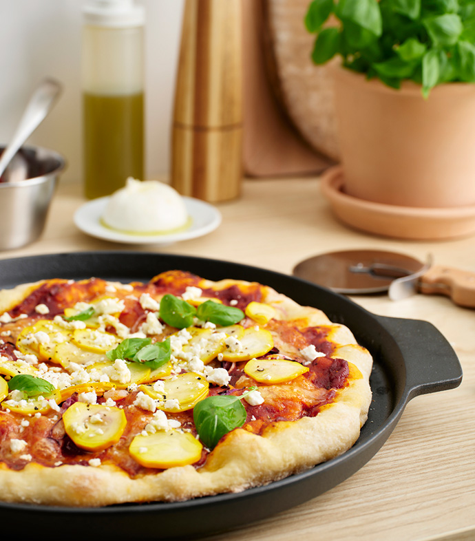 Pizza de calabacín y queso sobre sartén negra, con albahaca y utensilios de cocina al fondo.