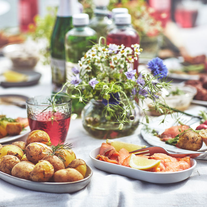  Outdoor table setting with roasted potatoes, salmon, meatballs, and a vase of wildflowers, with a sparkling water view in the background. 