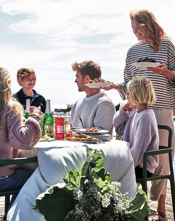  Group gathering at an outdoor table by the water, sharing a meal while a person in a striped shirt serves a plate of food.