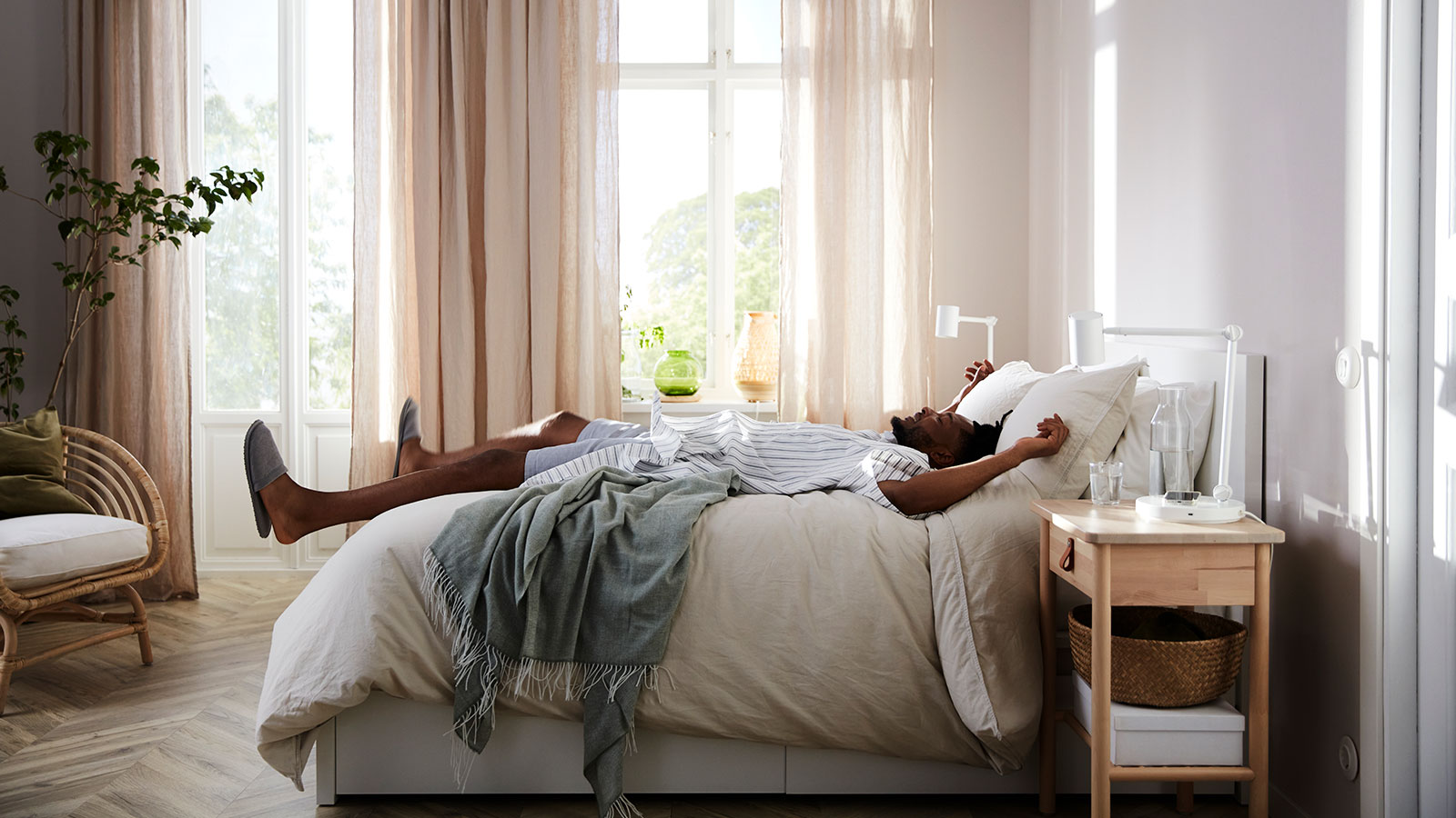 Image of a brightly lit bedroom with beige curtains in side view, with a man lying down as if he just 