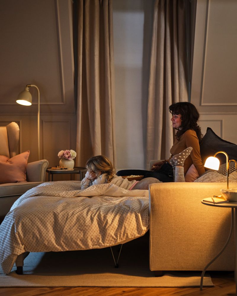 An adult and a child are on an EKHOLMA 2-seat sofabed which is configured as a bed near a STRANDMON wing chair and a lamp.