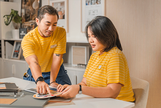 Two IKEA employees in yellow striped uniforms work together at a desk, looking at a circular color wheel and material samples.