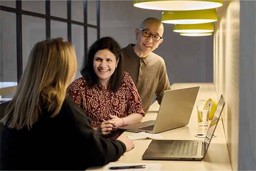Three colleagues having a cheerful discussion around laptops at a long desk under warm yellow pendant lights in a modern office space.