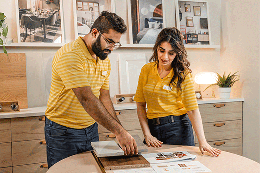 Two IKEA employees in yellow striped polo shirts stand at a table reviewing design documents and countertop material samples in a showroom.