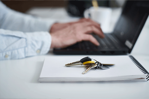A close-up focus on a set of keys resting on a blank notebook, with a person's hands blurred in the background typing on a laptop.