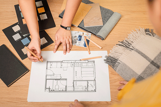 A top-down view of hands working on an architectural floor plan at a wooden desk, surrounded by color swatches, wood samples, fabric, and pencils.