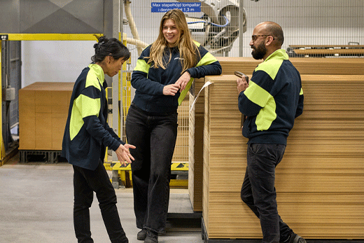 Three logistics workers in dark jackets with high-visibility yellow panels chat and laugh next to large stacks of flat-packed cardboard boxes in a warehouse.