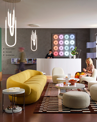 Un moderno vestíbulo de hotel con un sofá largo amarillo, mesas redondas blancas y una recepcionista en el mostrador bajo un panel de luces de colores.